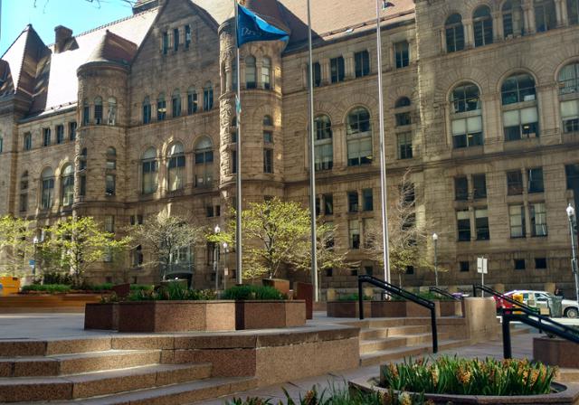 Allegheny County Courthouse, which is a large stone building, in the distance.  Sidewalk with lined with trees and yellow flowers in the foreground.