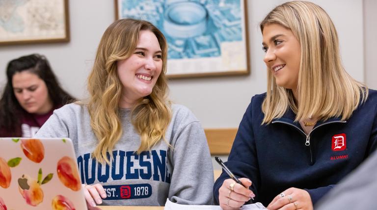 Two female students in front of a laptop, in pleasant discussion about a class assignment.