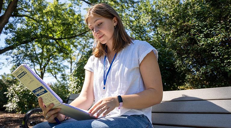 A student sits on an outdoor bench, reviewing a reporter's notebook in hand, with green trees visible in the background.