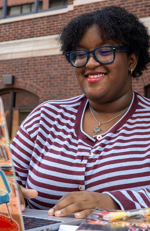 A student with short curly hair and blue glasses works on her laptop with textbooks nearby.