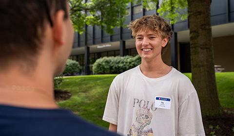 A student wearing a name badge smiles while talking with another student on Academic Walk, with Mellon Hall in the background.