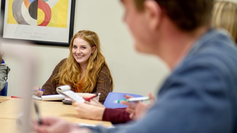 Female student smiling at camera from across a classroom desk