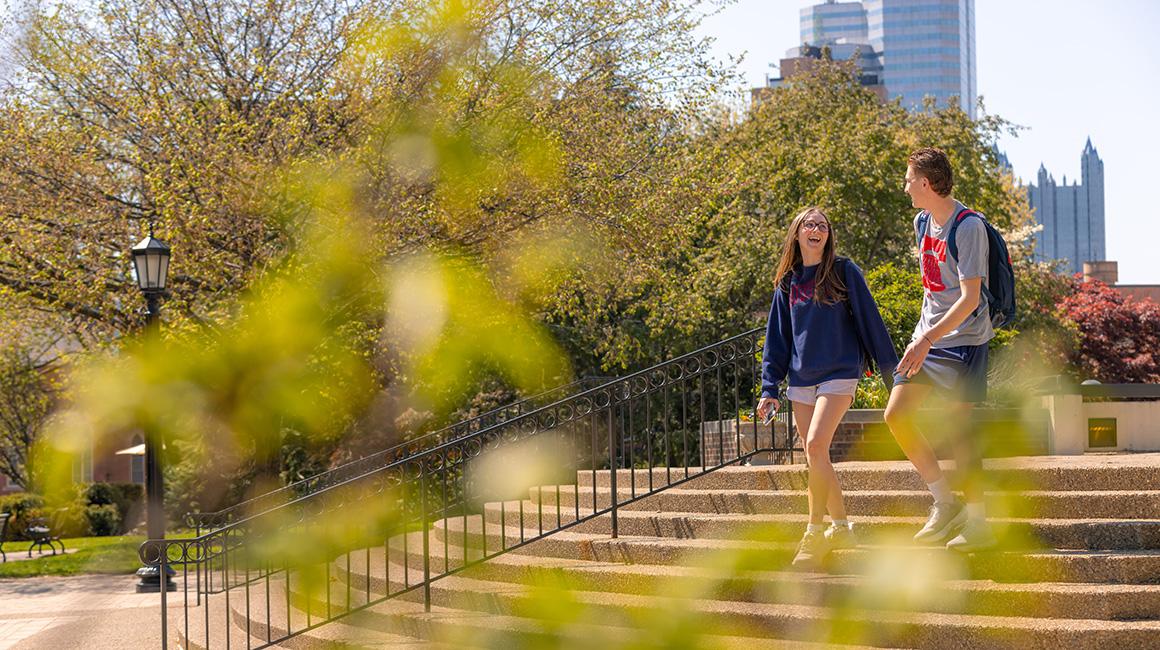 Two students walking down staircase with downtown Pittsburgh in background.