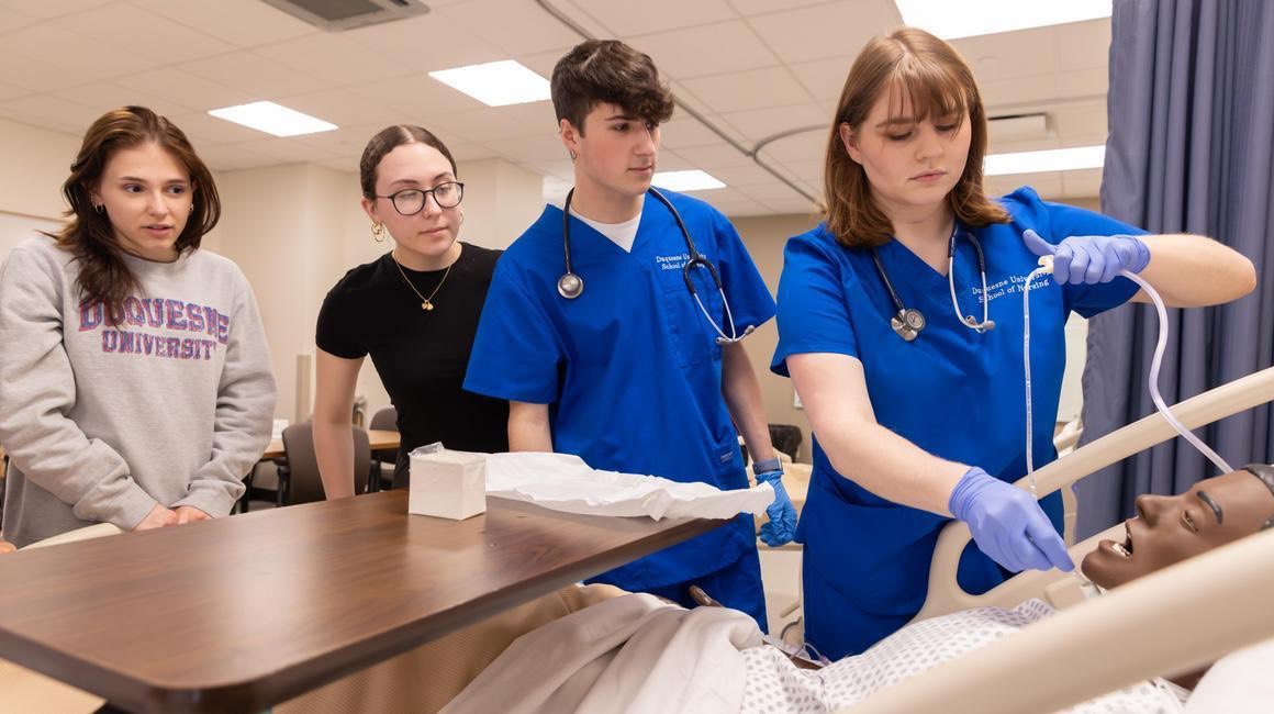 Students in scrubs practice clinical skills on a medical simulation mannequin in a nursing lab.