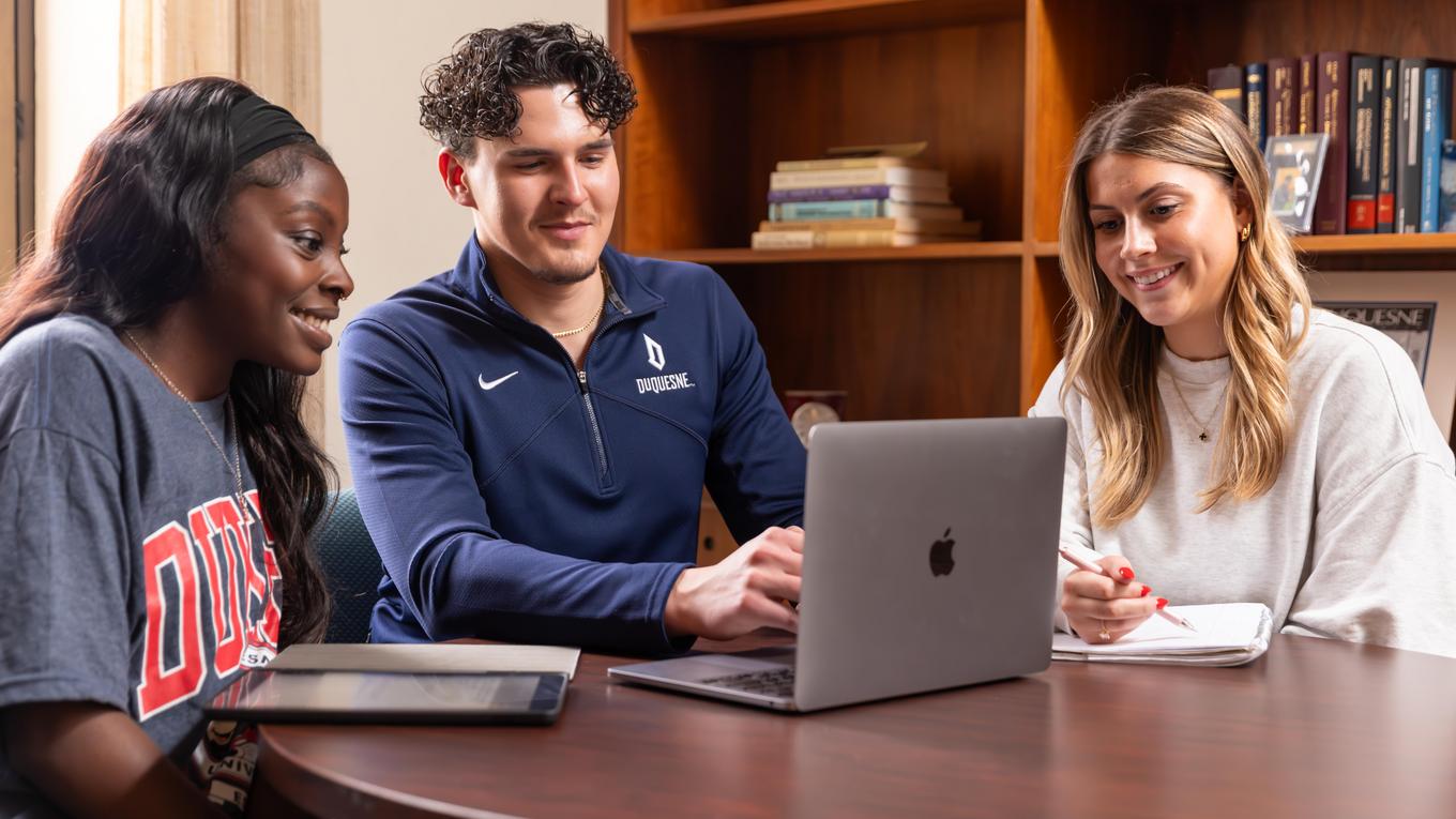 Two female students and one male student sitting around a laptop smiling