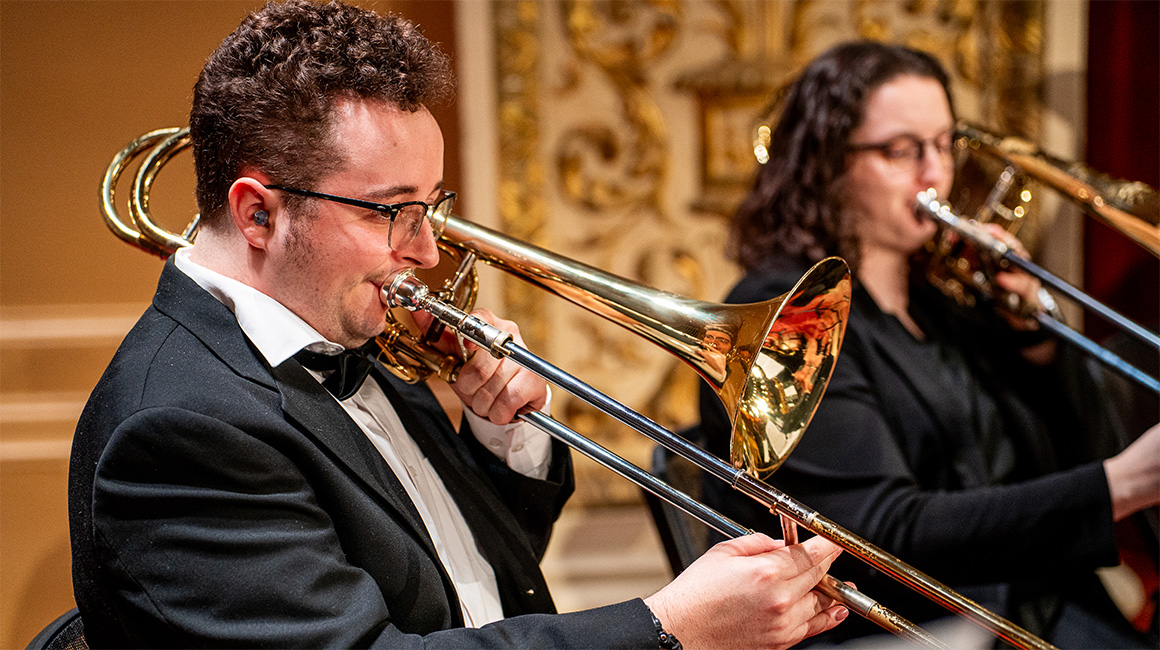 A trombonist in concert dress performs on stage. Another trombonist can be seen in the background.