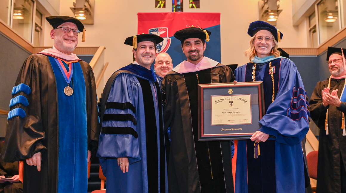 Four people pose in commencement regalia, and two hold a framed certificate.