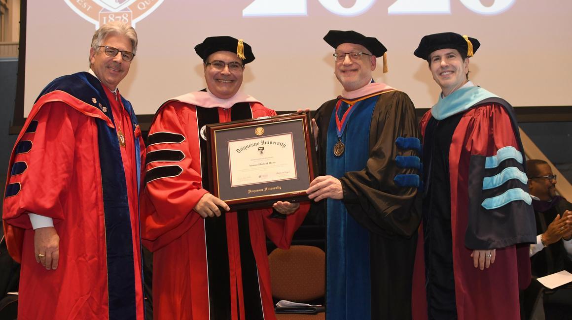 Four people in academic regalia pose for a photo. An award is being presented to one of them.