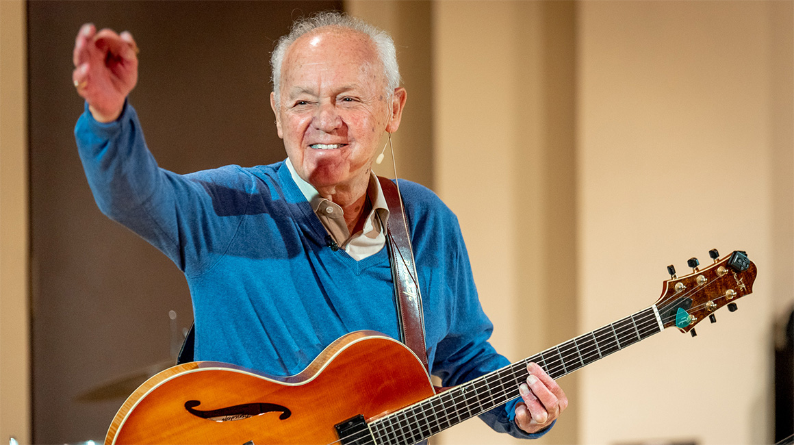 Joe Negri smiles and raises his hand while holding a guitar during a performance at Duquesne University's Mary Pappert School of Music.