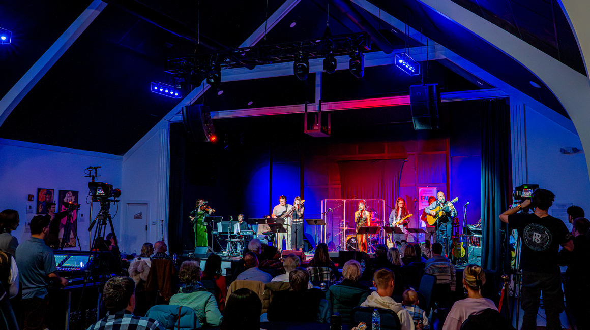 Wide view of a live concert with a full band performing on a covered stage under blue and purple lighting, an audience gathered in front, and multiple cameras capturing the performance.