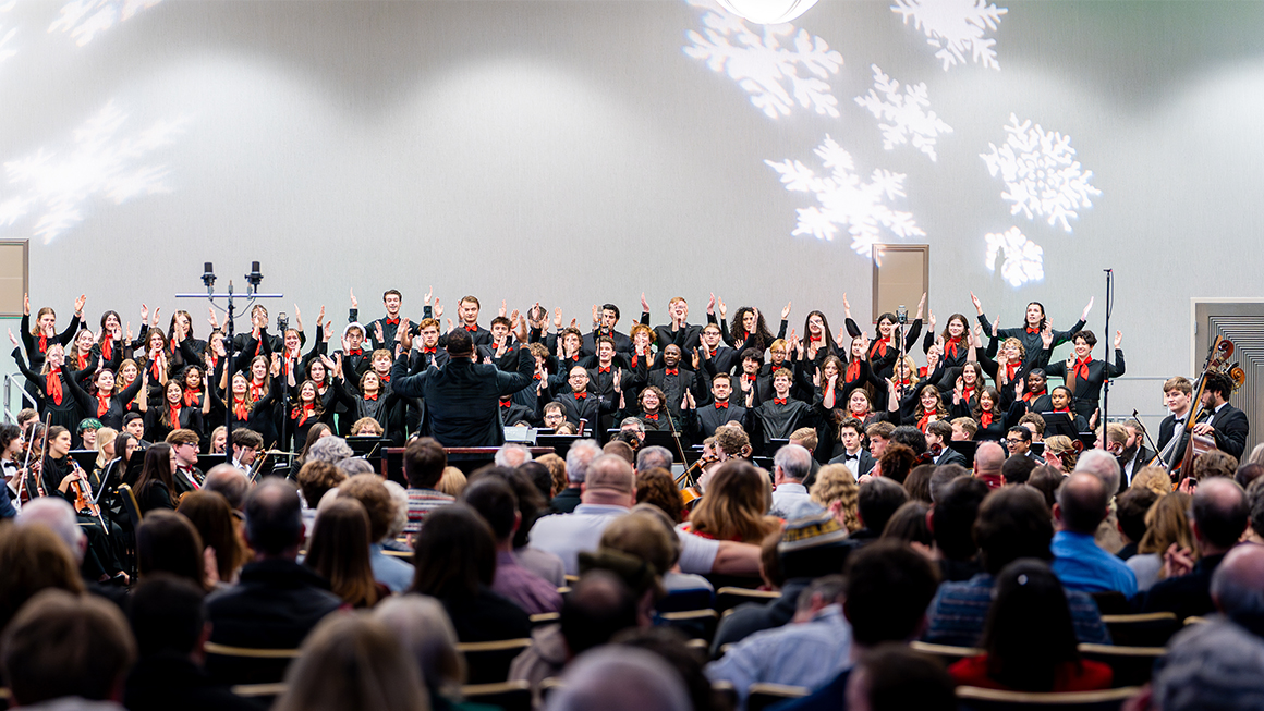 Wide shot of full choir and orchestra performing to a packed audience.