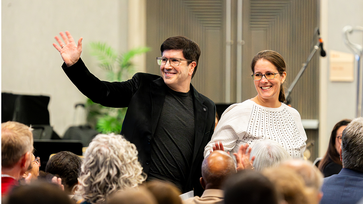 Two guests stand and wave to applauding audience inside concert hall.
