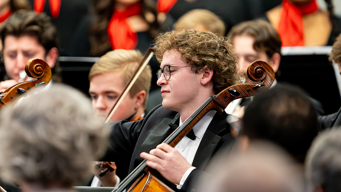 Cellist performs seated among string section with choir blurred behind.