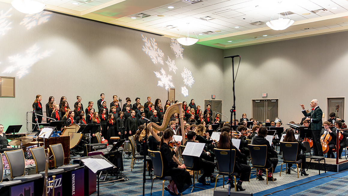 Wide view of orchestra, harp, and choir performing beneath projected snowflakes.