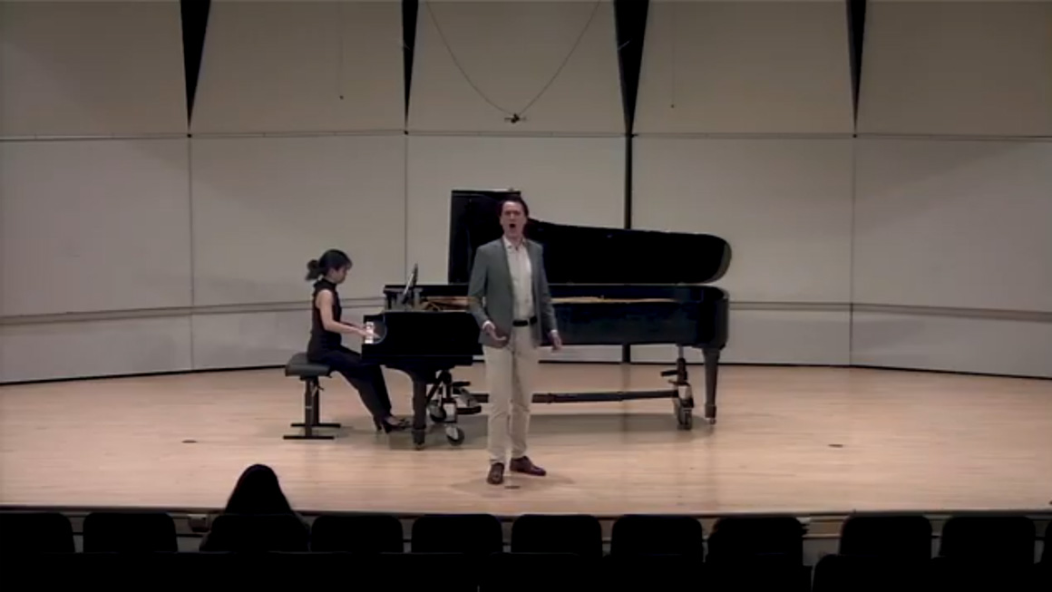 A person sings on stage with a pianist in the background.