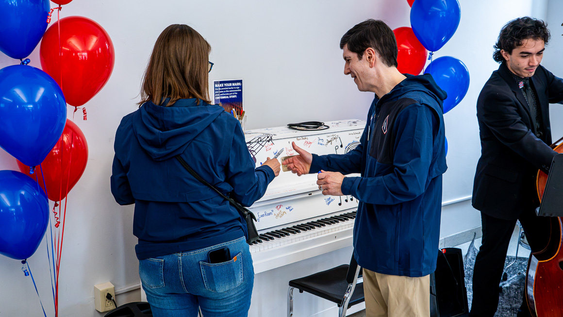 President David Dausey and his wife prepare to sign a white piano surrounded by red and blue balloons. A student bassist can be seen in the background.