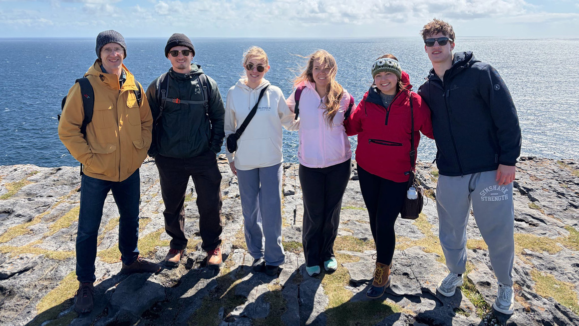 Six participants stand together on a rocky coastal overlook with the ocean stretching behind them. Dressed in jackets and hats against the wind, the group smiles for the camera beneath a bright sky reflected across the water.