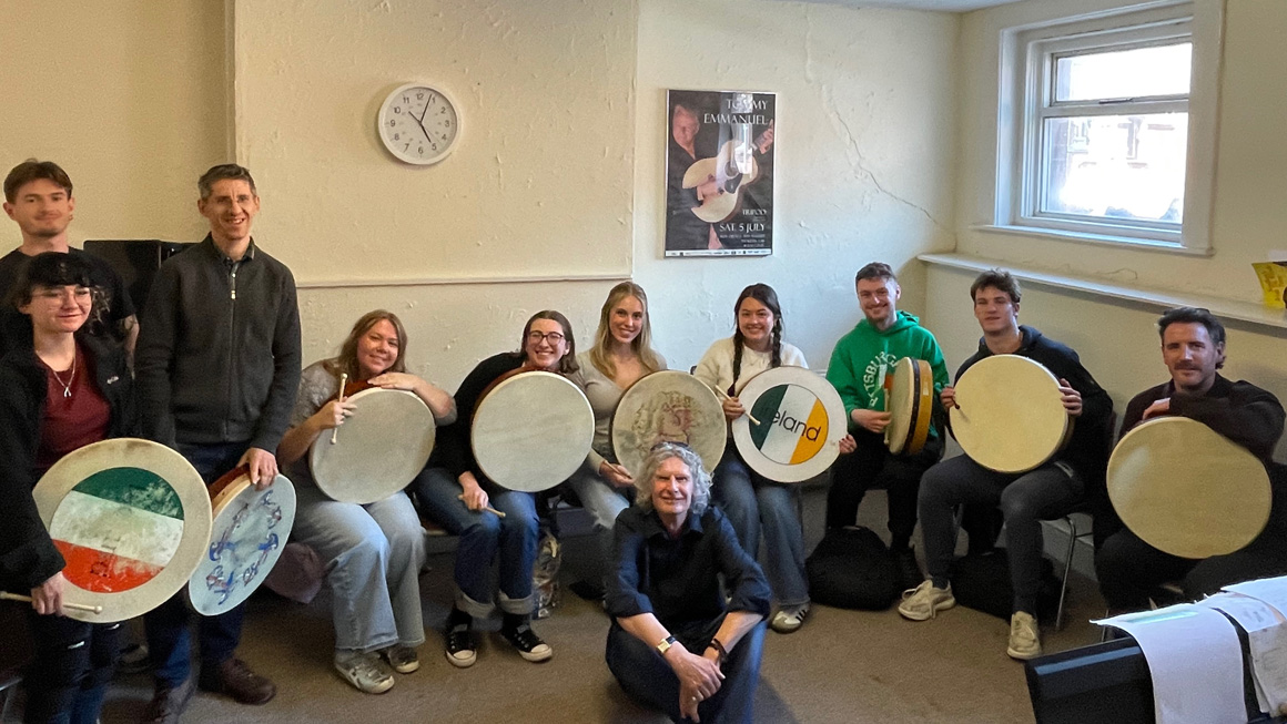 Students and instructors gather indoors holding traditional Irish frame drums during a hands-on music workshop. The group poses with their instruments while seated and standing around the classroom.