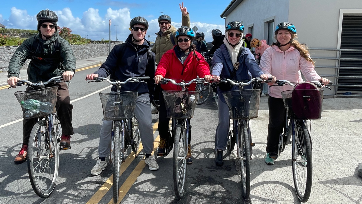 A group of students wearing bicycle helmets lines up on rental bikes before a ride through a coastal town. An instructor smiles behind them as the group prepares to set off under sunny skies.