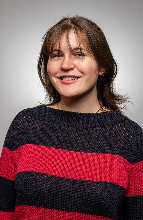 A person wearing a black and red striped sweater smiles in front of a gray background.