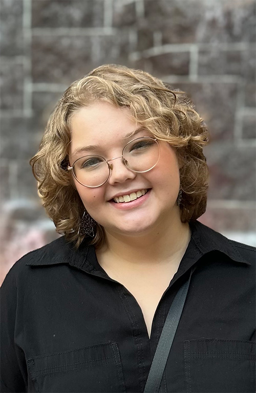 A person with curly blonde hair and glasses smiles in front of a brick background.