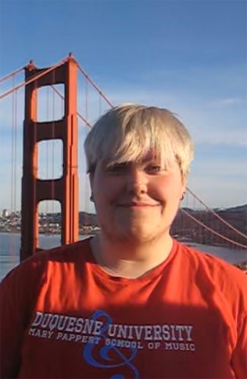 A person with blonde hair wearing a red Mary Pappert School of Music t-shirt stands in front of the Golden Gate Bridge.