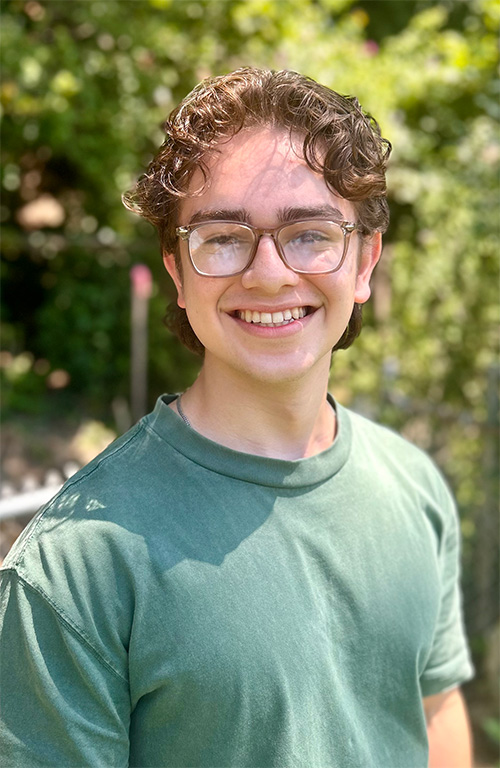 A person wearing a green shirt and glasses smiles for a portrait in a wooded area.