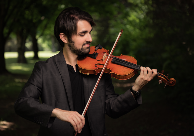 Ryan Joseph plays a violin in front of a grove of trees.