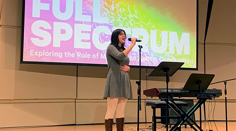 A person sings into a microphone on stage next to a keyboard and in front of a projection with the Full Spectrum logo.