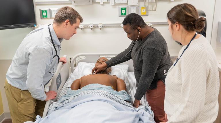 A woman using a stethoscope to check on a simulation mannequin. A man and woman stand around her and watch. They are in a clinical setting.