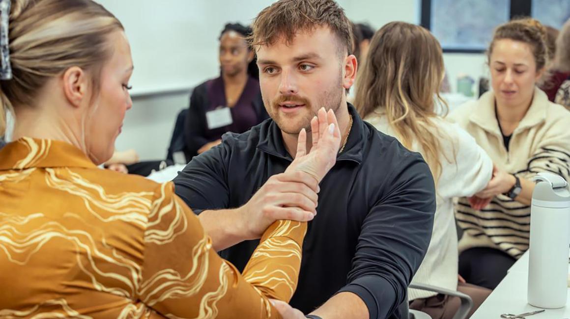 A male student examining a female students arm. There are groups of two students around them doing the same thing.
