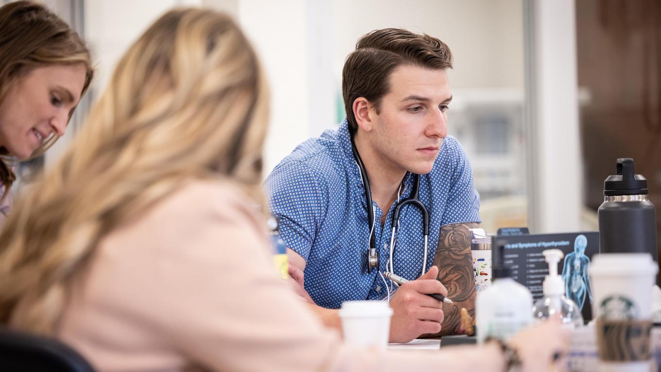 Student with stethoscope around neck talking to a group of peers at a table. 