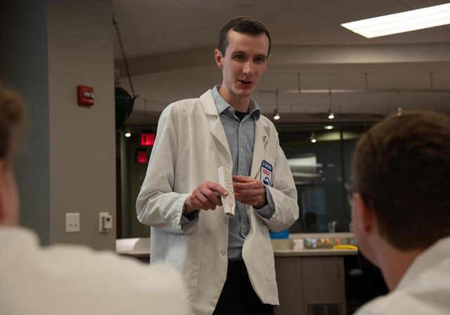 student in lab coat speaking to other students