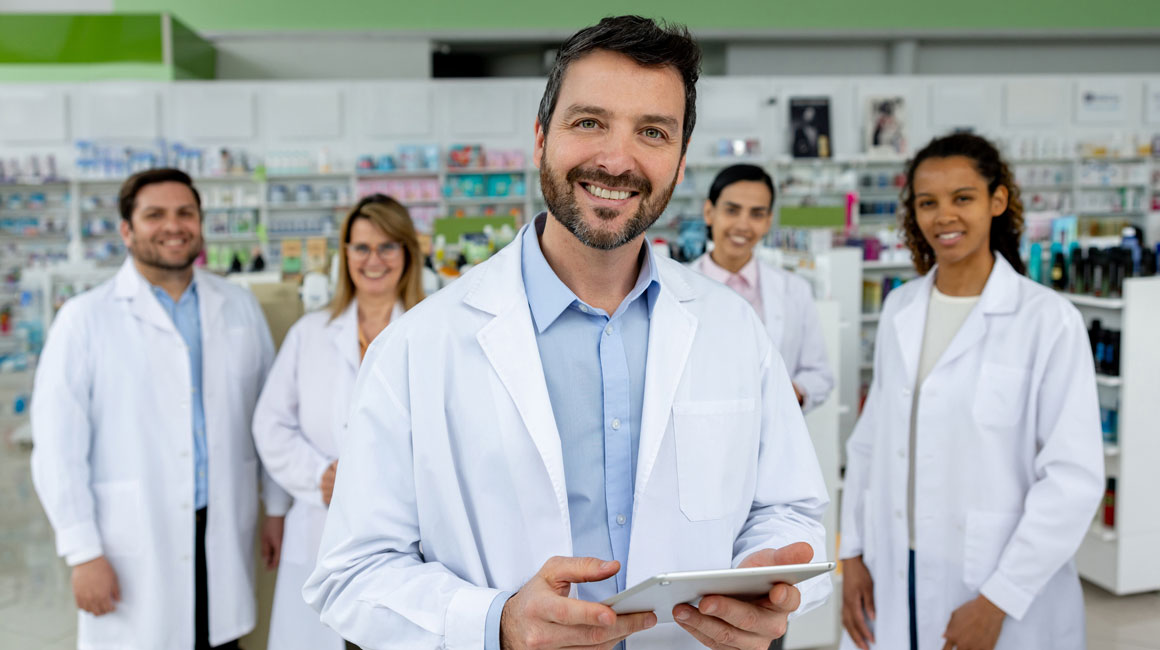 male pharmacist smiling at camera with additional pharmacists standing behind him