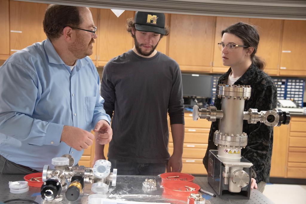 two students and a professor in front of a piece of equipment