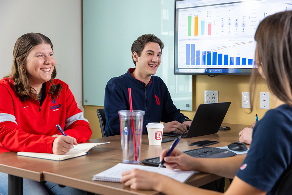 Three Duquesne students working together at a table with a laptop and bar graphs in the background