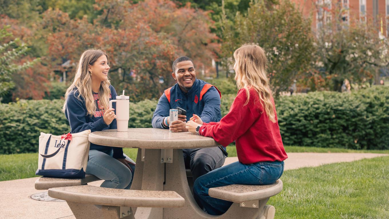 Duquesne students talking at a table outside on campus with greenery in the background