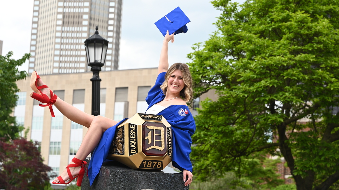 A DU graduate sits on the Red Ring holding her graduation cap high.