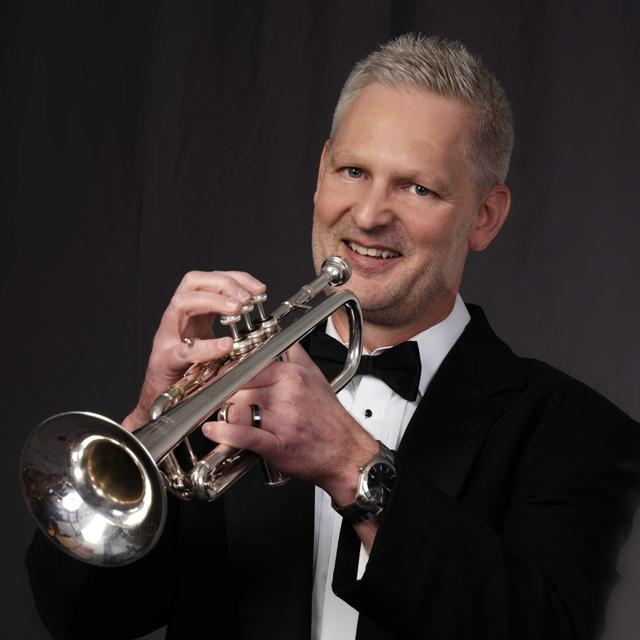 A person in a tuxedo holds trumpet in front of black background.