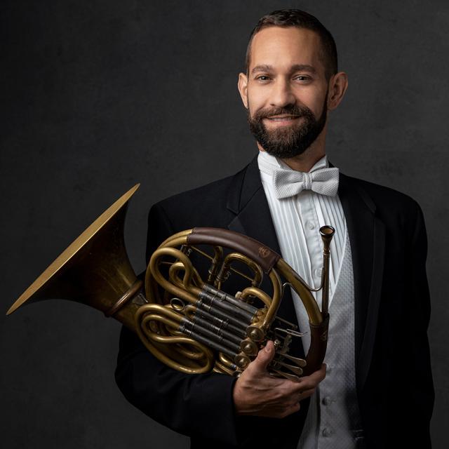 Mark Houghton wears a tuxedo and holds a French horn.