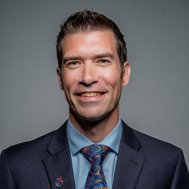 A person wearing a suit and tie smiles in front of a neutral background.