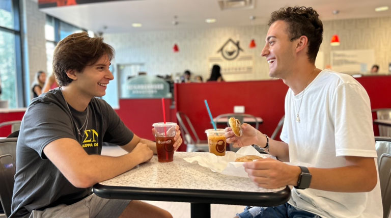 Two students sit at a table enjoying drinks and a sandwich from The Neighborhood Roasters.