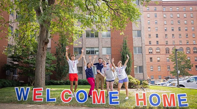 Students jumping with joy behind a red and blue welcome home sign