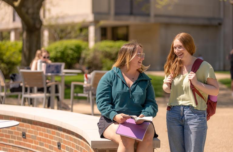 Two female students are talking to each other, smiling, while hanging out on campus on a sunny day.