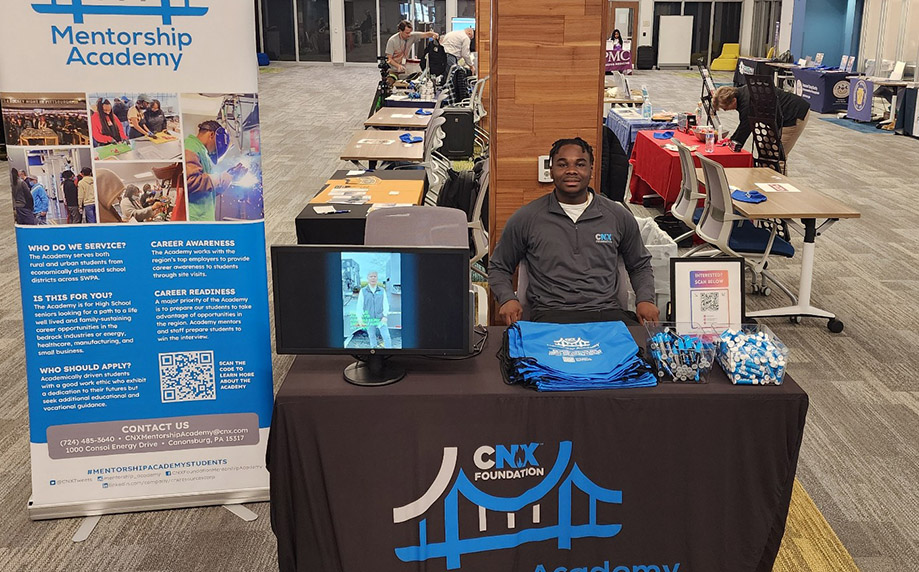 Mwango Kasha sits behind a table representing CNX Mentorship Academy at a career fair