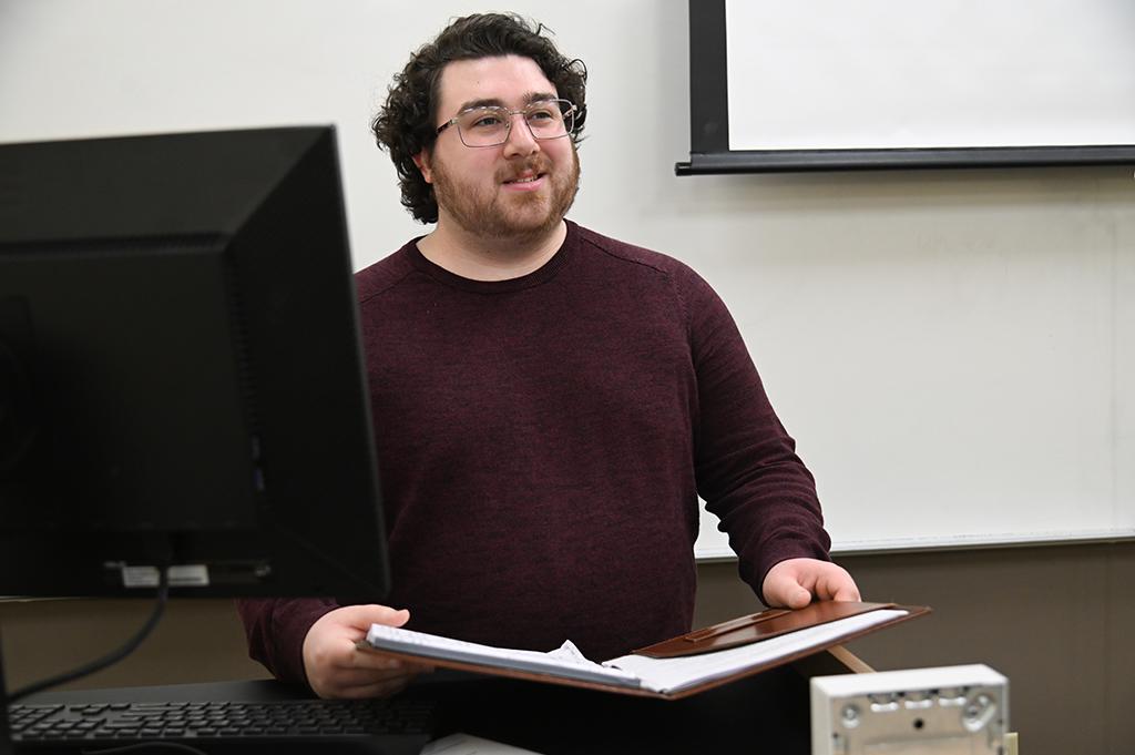 Nick Freilino speaks in front of a classroom holding a brown folder and notebook