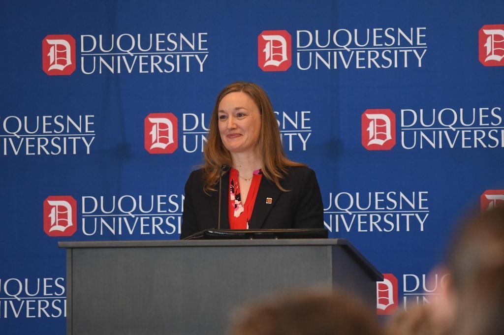 Dr. Sarah Ewing addresses a crowd from a podium in front of a Duquesne University backdrop. 
