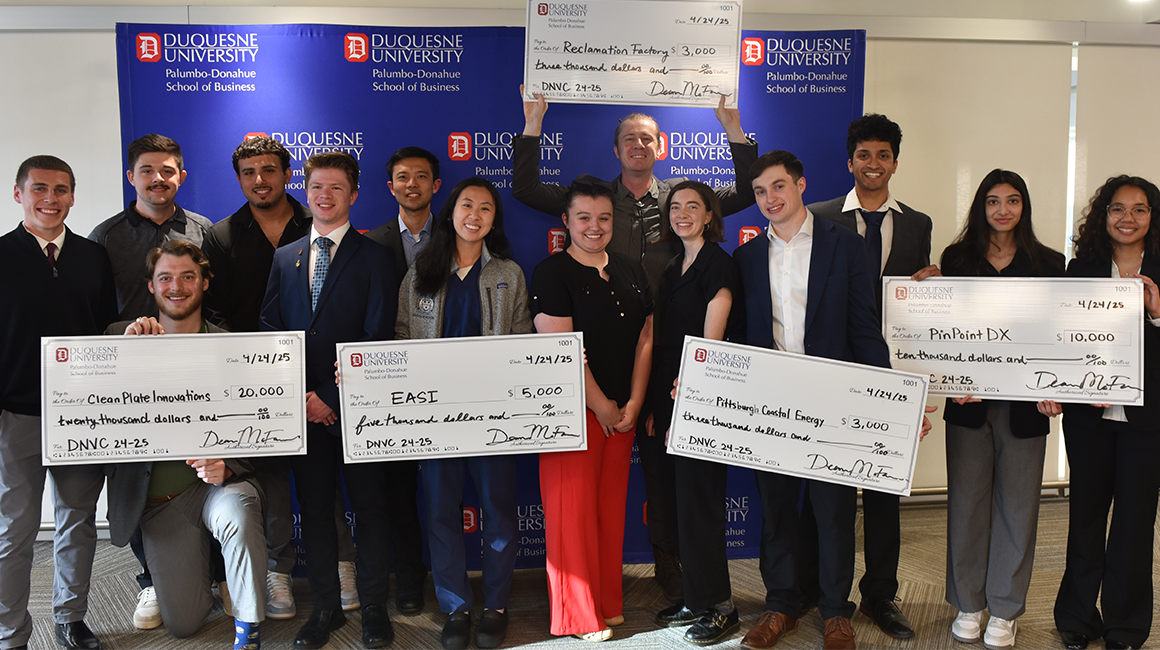 Winners of the 2024 - 2025 Duquesne New Venture Challenge stand in front of a Duquesne University backdrop holding large checks.