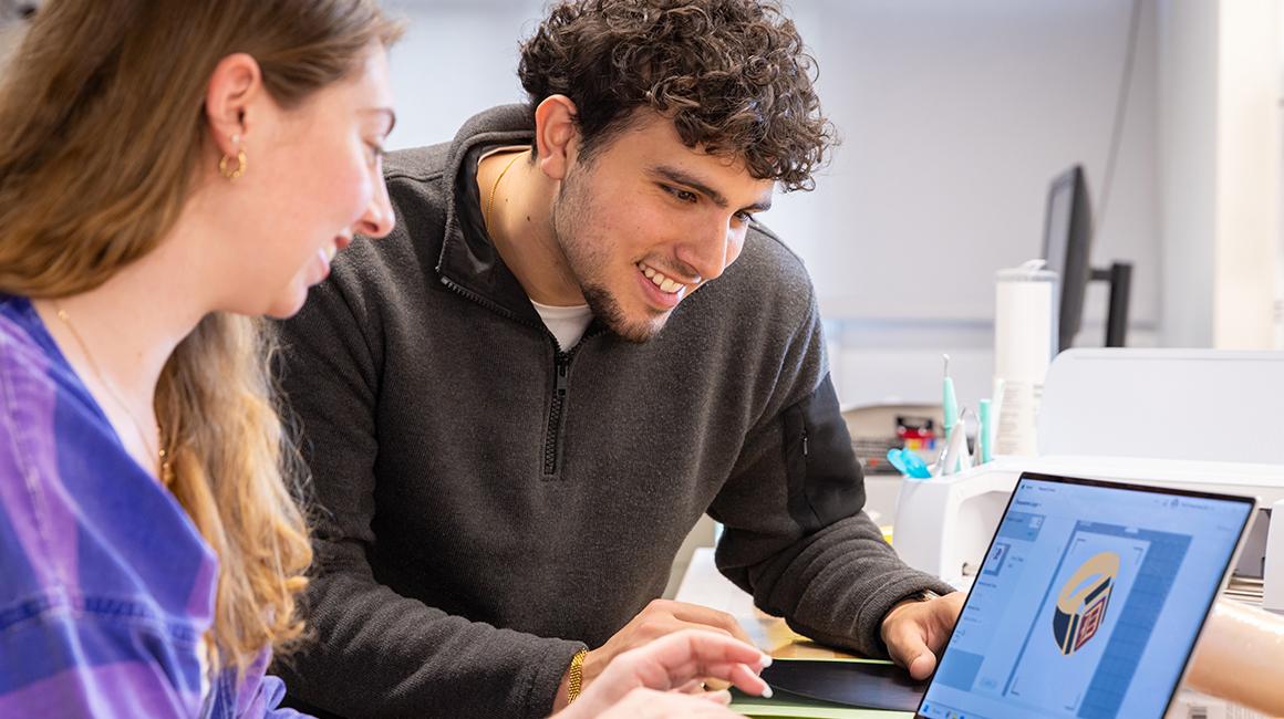 Two students look at a laptop screen featuring a graphic of the Duquesne ring.