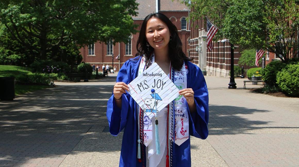 Joy Chen holds her graduation cap and stands on Academic Walk beneath a Commencement 2025 banner